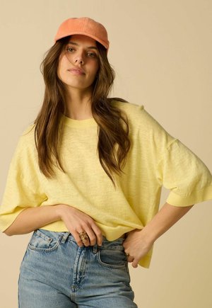 Jeune femme aux longs cheveux bruns portant une casquette orange, un haut jaune ample, un jean bleu taille haute et des bagues en or, posant avec une main dans la poche.