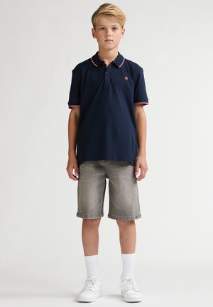 Marineblauw poloshirt met rode bies, gecombineerd met lichtgrijze denim shorts. Witte sneakers en kousen tot net onder de knie maken de outfit compleet.