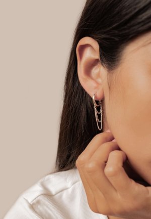 Boucles d'oreilles créoles argentées avec un design à chaînes superposées, présentant une finition lisse et des maillons délicats, portées près de l'oreille d'une personne.