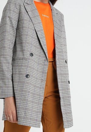 Vrouw draagt een zwart-wit geruit double-breasted blazer over een oranje shirt met de tekst "Journey" en een bruine broek.