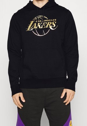 Person iført sort Los Angeles Lakers hættetrøje med guldlogo og sorte bukser med lilla detaljer og Lakers-patch.