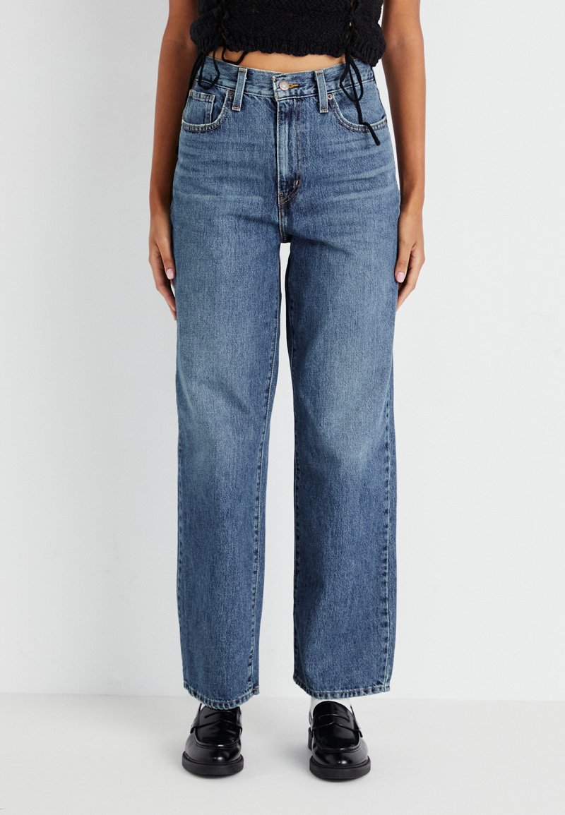 Personne portant un jean bleu taille haute à jambes droites avec des mocassins noirs et un haut court noir sur un fond blanc uni.