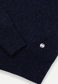 Námornícko modrý sveter s textúrovaným pletením. Obsahuje rebrované manžety a malú kovovú nálepku. Mäkkučký materiál so subtílnymi zelenými bodkami.