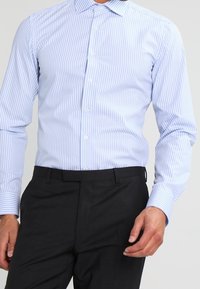 Man in een lichtblauw en witte verticaal gestreepte overhemdbijl, ingestopt in een zwarte pantalon, staand met één hand ontspannen.