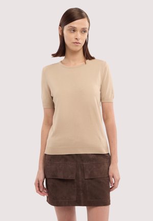 Donna che indossa una maglia beige a maniche corte in maglia e una minigonna in camoscio marrone con grandi tasche frontali, in piedi contro uno sfondo semplice.