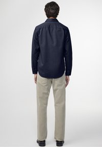 Camicia navy a maniche lunghe con polsini a bottone, cucitura dritta sul retro, abbinata a pantaloni di un beige chiaro e scarpe nere, su uno sfondo neutro.