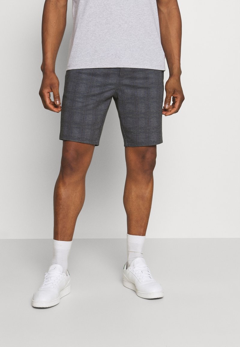 Homme portant un short gris à carreaux, des chaussettes blanches, des baskets blanches et un t-shirt gris clair, debout sur un fond uni.