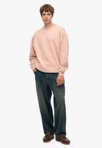 Felpa a girocollo rosa chiaro, vestibilità comoda, indossata con jeans in denim blu scuro. Scarpe in pelle marrone con lacci e suole texture.