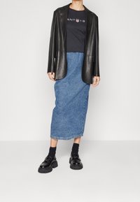 Person iført en sort læder-blazer, sort trykt t-shirt, blå denim midi-skjørt, sorte sokker og kraftige sorte loafers, der står indendørs.