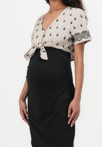 Vestido premamá con parte inferior negra ajustada y parte superior beige con nudo delantero, decorado con pequeños patrones negros y detalles tribales.