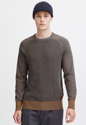 Pull en tricot marron avec un motif texturé, col rond côtelé et ourlet marron contrastant. Un petit patch de marque est cousu en bas à gauche.