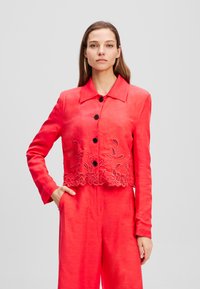Rote, verkürzte Jacke mit langen Ärmeln, großem Kragen, Knopfverschluss und floralem Stickereimuster am Saum, aus glattem, leichtem Stoff gefertigt.