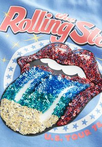 Sininen paita, jossa on paljeteilla varustettu kuva huulista ja kielestä punaisista, sinisistä, vihreistä ja kultaisista paljeteista, sekä teksti "The Rolling Stones".
