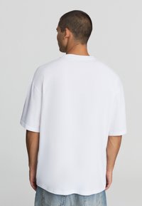 Biała, oversize koszulka z krótkim rękawem i okrągłym dekoltem, wykonana z materiału bawełnianego, z gładką fakturą i prostym wykończeniem. Brak widocznej marki.