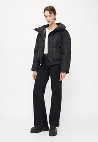 Crna puffer jakna s odvojivim kapuljačom, zatvaranjem na zip, i šivenim detaljima, nosi se preko bijele majice i u kombinaciji s crnim trapericama visokog struka.