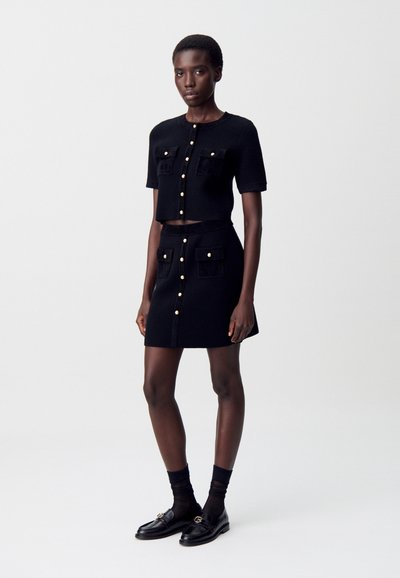 Femme portant un crop top noir à manches courtes et boutons, accompagné d'une jupe assortie ornée de boutons dorés et de poches, des chaussettes noires et des mocassins.