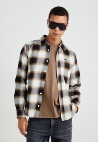 Camisa de cuadros en tonos blanco, azul y marrón con diseño de botones; llevada sobre una camiseta beige; presenta un cuello y mangas largas.