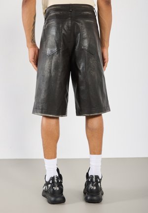 Personne portant un short en cuir noir jusqu'aux genoux, des chaussettes blanches montantes et des baskets noires, debout devant un fond clair uni.