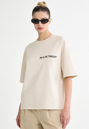 Femme aux cheveux blonds attachés en chignon, portant des lunettes de soleil noires, un t-shirt beige ample avec le texte "YOU & ME TONIGHT" et un pantalon beige assorti.