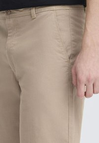 Pantalones de algodón beige con una textura suave, que cuentan con bolsillos frontales y detalles de costura sutiles. Vista en primer plano que muestra la cintura y la colocación de la mano.
