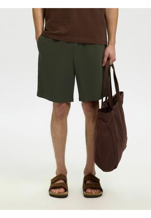 Personne portant un short vert foncé, un haut marron et des sandales marron tenant un grand sac fourre-tout marron, debout devant un fond uni.