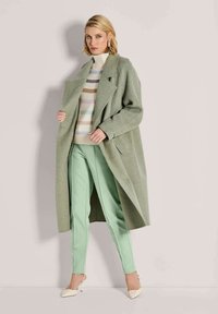 Vrouw in een pastelgroene lange jas, gestreepte coltrui, mintgroene broek en crèmekleurige puntige hakken, staand tegen een beige muur.
