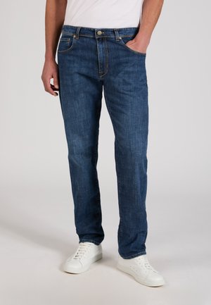 Man wearing dark blue straight-leg jeans with white sneakers, standing with one hand in pocket against plain background.