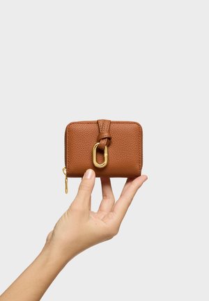 Dłoń trzymająca mały brązowy portfel z teksturowanej skóry z złotym zamkiem błyskawicznym i dekoracyjnym złotym zapięciem na jasnym tle.