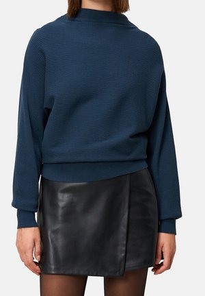 Personne portant un pull côtelé bleu foncé à manches longues et une mini-jupe en cuir noire avec des collants.