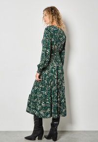 Groene midi-jurk met een bloemenpatroon, gesmokte taille, lange mouwen en een gelaagde rok. Draagt met kniehoge zwarte laarzen.