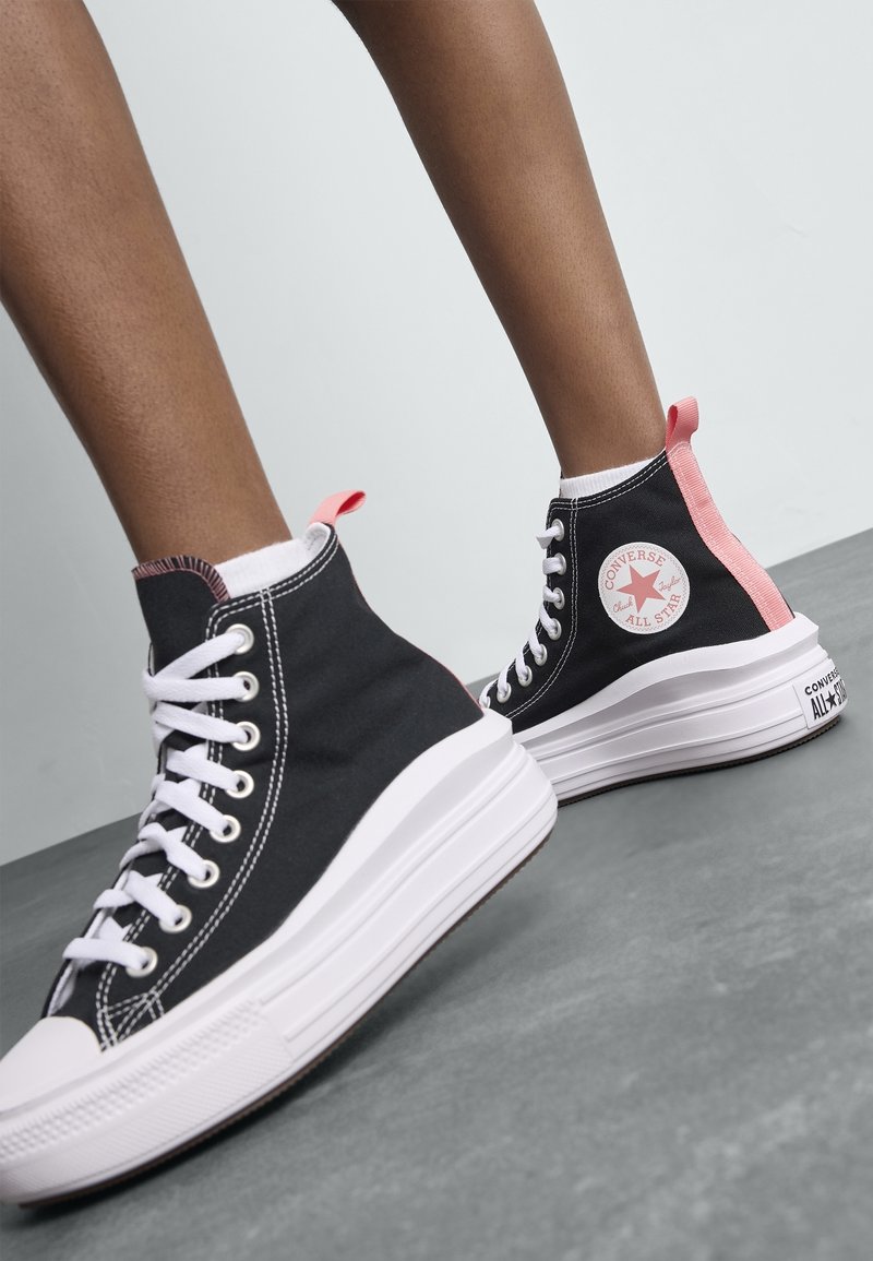 Jambes portant des baskets montantes Converse All Star noires avec des lacets blancs, des semelles blanches épaisses et des languettes de talon roses sur un sol gris.
