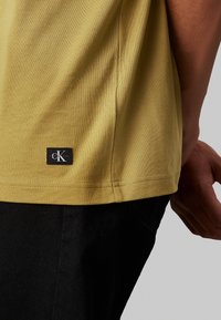 T-shirt khaki a maniche corte realizzato in morbido cotone, caratterizzato da un patch logo CK nero vicino all'orlo, abbinato a jeans in denim scuri.