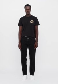 Tricou negru cu un logo aurit pe piept, asortat cu blugi negri cu cusături albe și adidași albi. Design simplu, ajustat.