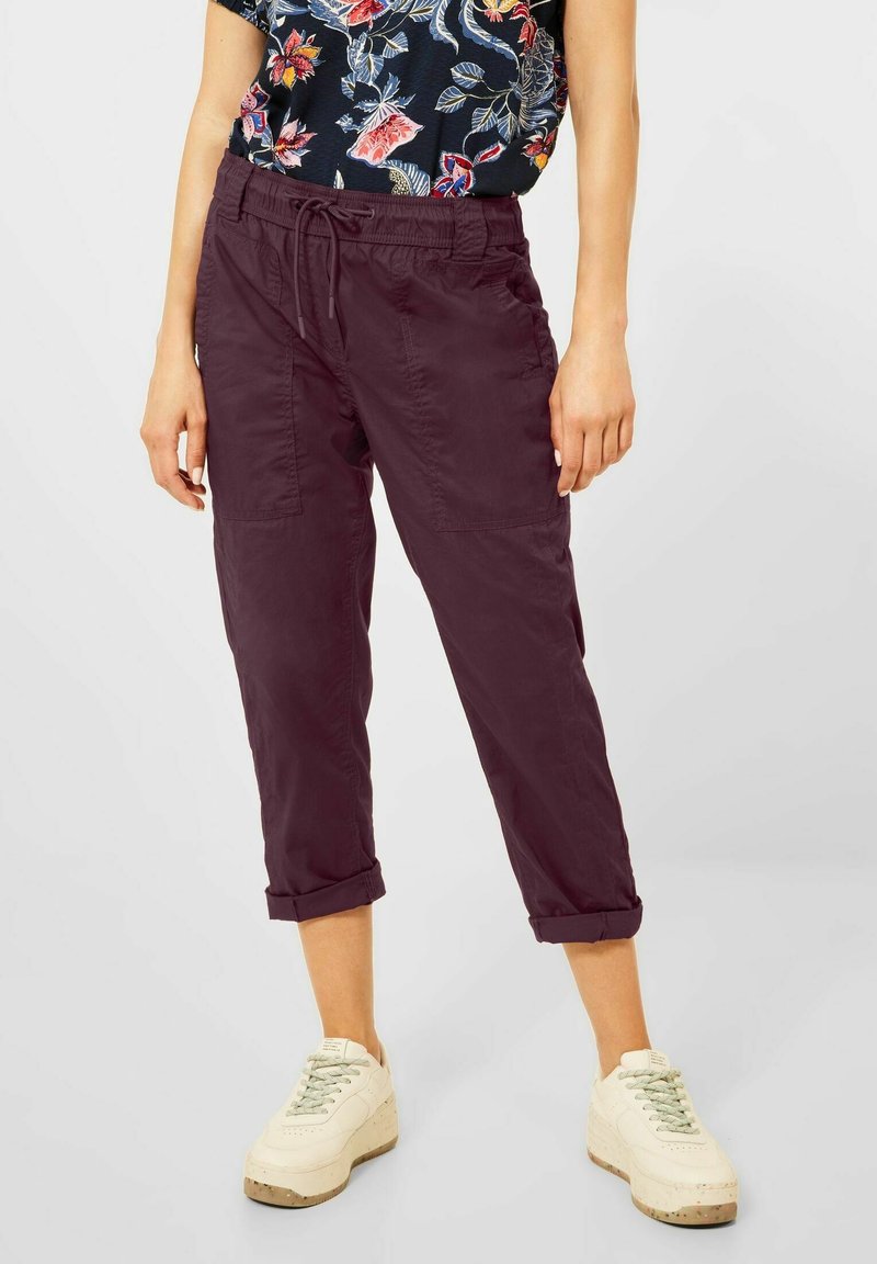 Personne portant un pantalon violet foncé retroussé, une chemise fleurie bleu marine et des baskets blanches, debout sur un sol gris clair sur fond uni.