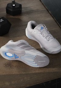 Ζευγάρι λευκά και γκρι παπούτσια Adidas για τρέξιμο με μπλε λεπτομέρειες, το ένα δείχνει τη λάστιχη σόλα Continental, σε ξύλινο πάτωμα κοντά σε δύο μαύρα αλτήρες.