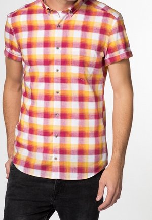 Mężczyzna ubrany w koszulę na guziki z krótkim rękawem w czerwono-żółto-białą kratę oraz czarne spodnie, stojący na białym tle.