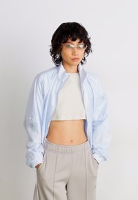 Persona con capelli ricci e scuri che indossa occhiali trasparenti, un giubbotto azzurro chiaro sopra una maglietta bianca corta e pantaloni beige con le mani in tasca.