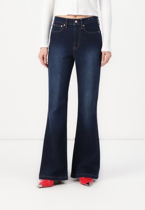 Dark blue flared jeans with stitched back pockets, copper-tone hardware, and sleek texture. Worn with red pointed-toe shoes.
