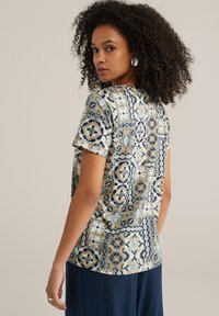 Gaišzils īsām piedurknēm tops ar sarežģītiem ģeometriskiem rakstiem krēmkrāsā, jūras zila un brūnā krāsā, ar brīvu piegriezumu un mīkstu kokvilnas materiālu.