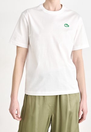 Personne portant un t-shirt blanc uni à manches courtes avec un petit logo crocodile vert sur la poitrine et un pantalon ample vert olive, debout devant un fond blanc.