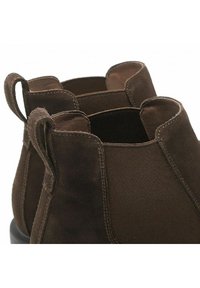 Stivali ankle in suede marrone con pannelli laterali elastici, tomaia tagliata in diagonale e piccole linguette per la trazione. Superficie testurizzata con dettagli di cucitura a contrasto.