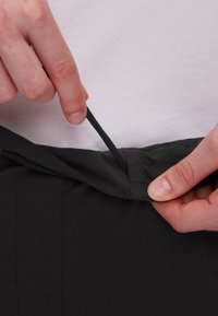 Schwarze Hosen mit einem elastischen Bund, bei dem eine Kordelzugverstellung vorgenommen wird. Der Stoff wirkt glatt und leicht, ideal für Freizeitkleidung.
