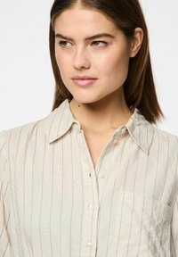 Camicia colletto beige chiaro con strisce blu verticali, chiusura frontale a bottoni e tasca sul lato sinistro, realizzata in un tessuto texture.