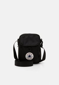 Svart tygväska med axelrem, klaffstängning, justerbar rem och en vit cirkelformad lapp med en stjärna och varumärkeslogotyp.