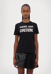 Mustmust valge ja metallhõbedase bold "Versace Jeans Couture" logoga must T-särk, kombineeritud musta volditud seeliku ja nahkdetailidega.