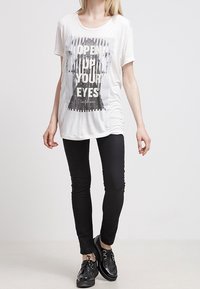 Vit bomullströja med ett grafiskt tryck som visar texten "ÖPPNA DINA ÖGON." Kombinerad med svarta skinnybyxor och texturerade svarta skor.