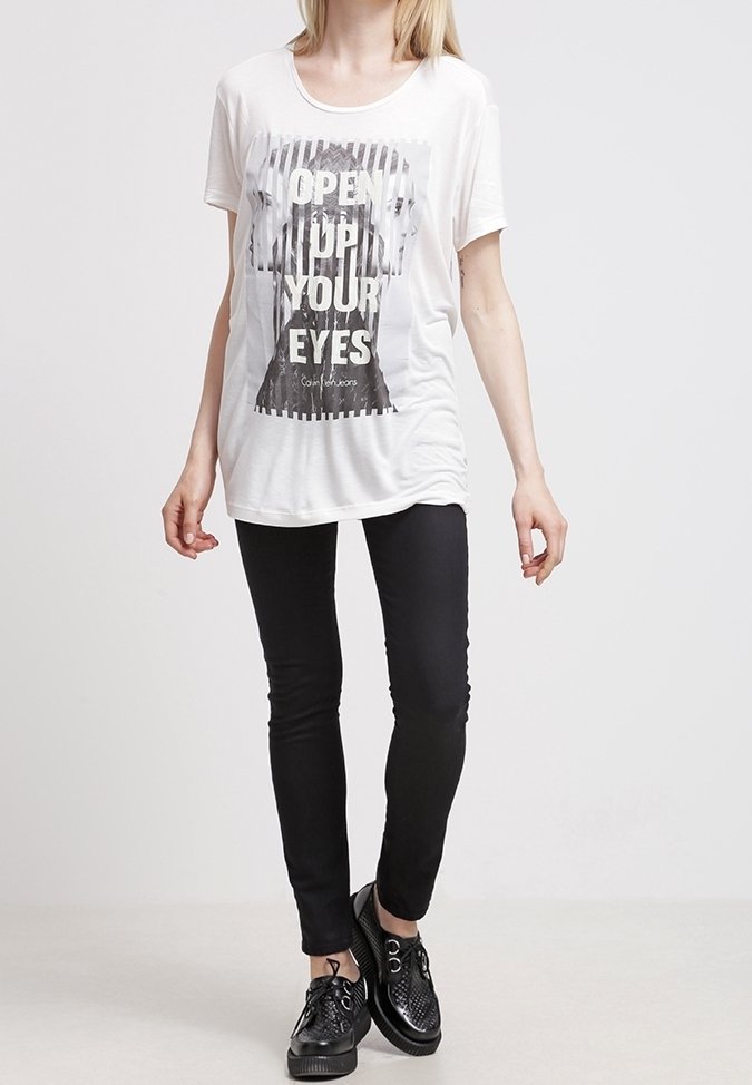 Vit bomullströja med ett grafiskt tryck som visar texten "ÖPPNA DINA ÖGON." Kombinerad med svarta skinnybyxor och texturerade svarta skor.