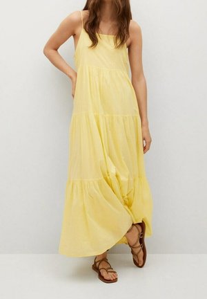 Kobieta ubrana w płynącą żółtą sukienkę maxi bez rękawów i brązowe płaskie sandały na paski, stoi przy gładkiej jasnej ścianie.