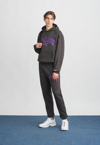 Σκούρα γκρι φούτερ με κουκούλα με μωβ λογότυπο "Reebok", συνδυασμένο με σκούρο γκρι φόρμα. Και τα δύο ρούχα είναι κατασκευασμένα από απαλό, υφασμα με υφή. Λευκά αθλητικά παπούτσια.