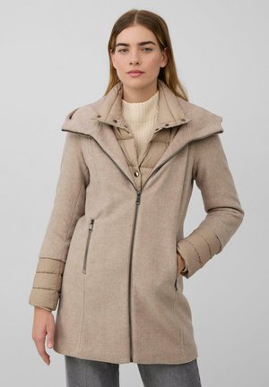 Kobieta w beżowym płaszczu z wełnianej mieszanki z pikowaną podszewką i mankietami, stojąca na tle jednolitego jasnoszarego tła.
